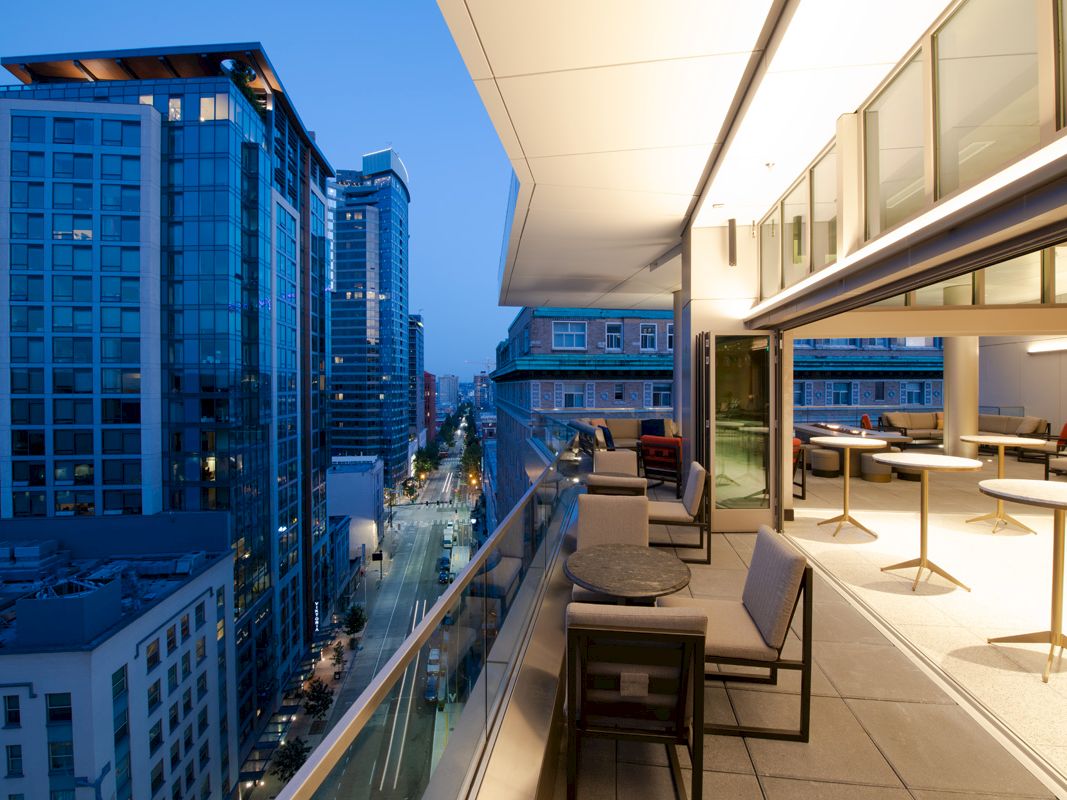 A modern urban balcony at dusk featuring seating and tables overlooks a city street lined with tall glass buildings.