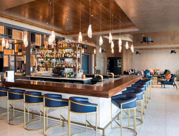 The image shows a modern bar with a wooden counter, blue bar stools, hanging lights, and a well-stocked bar shelf. There's seating in the background.