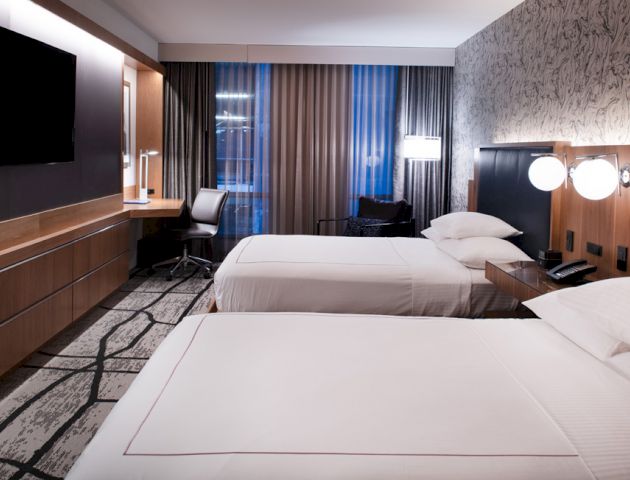 A modern hotel room with two beds, a wall-mounted TV, a desk, lamps, and patterned wallpaper, featuring sleek and clean decor.