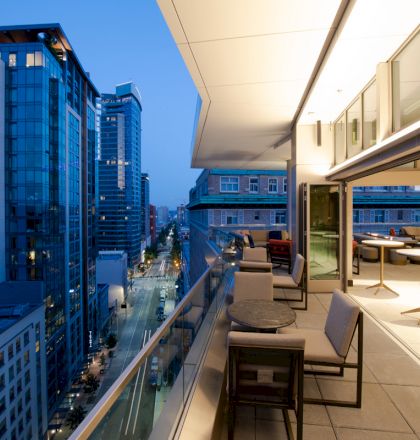 A modern balcony overlooking a cityscape with tall buildings and skyscrapers at twilight, featuring outdoor seating and tables.