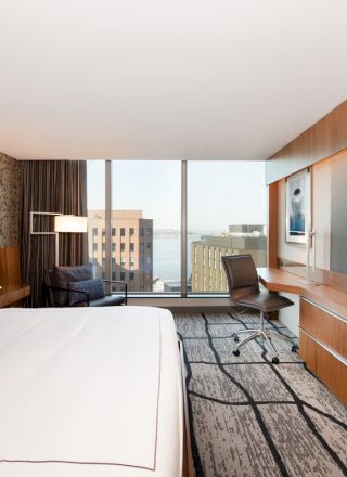 A modern hotel room with a king-size bed, desk, chair, large TV, and a window with a city view. The decor is contemporary and clean.