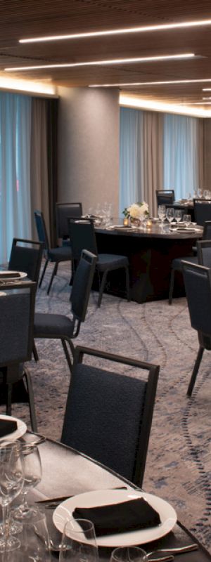 The image shows a banquet hall set up for an event with round tables, black chairs, and elegant place settings, including glasses and napkins.