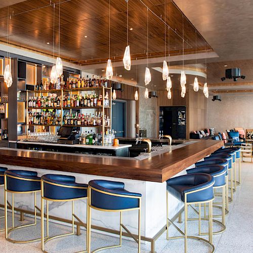 The image shows a modern bar with blue cushioned stools, a well-stocked liquor shelf, pendant lights, and a comfortable seating area in the background.