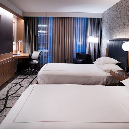 The image shows a modern hotel room with two neatly made beds, a wall-mounted TV, a work desk, and elegant lighting, creating a cozy atmosphere.
