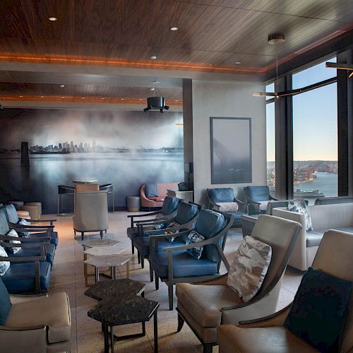 The image shows a modern lounge with comfortable seating, a large mural, and a stunning view of a waterfront through floor-to-ceiling windows.