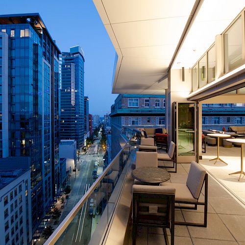 The image shows a modern cityscape at dusk, with a view from a balcony overlooking tall buildings and a street below. The balcony has seating and tables.