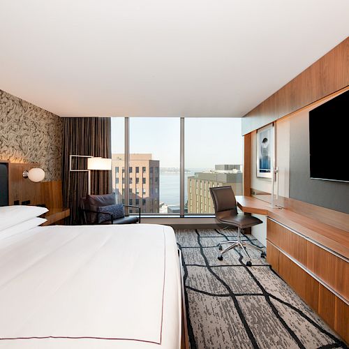 A modern hotel room featuring a large bed, wall-mounted TV, desk, chair, and floor-to-ceiling windows with a cityscape view.
