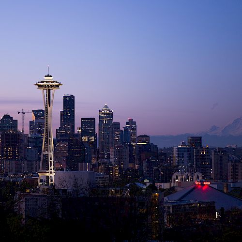 The image showcases a city skyline at dusk with a prominent tower, likely Seattle's Space Needle, and a mountain in the distance under a purple sky.