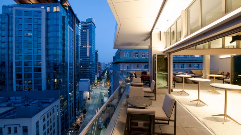 Image of a modern cityscape with tall buildings, viewed from a balcony with outdoor seating and tables at dusk, lights illuminating the scene.