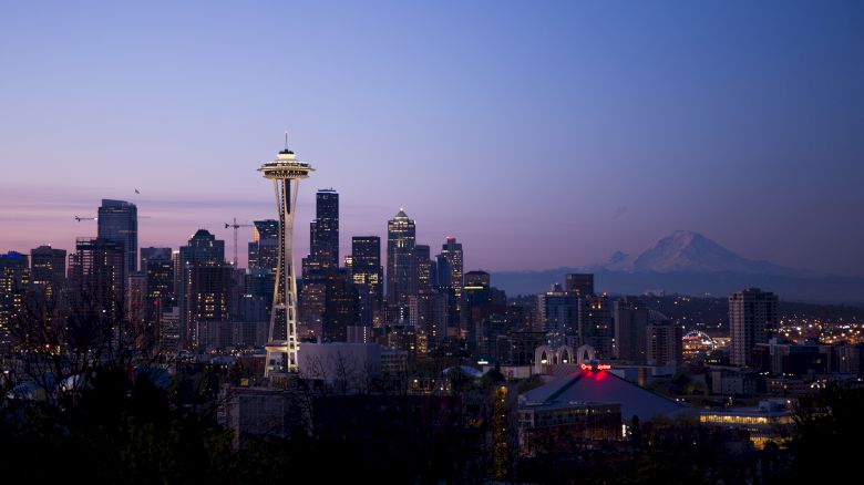This image depicts the Seattle skyline at dusk, featuring the Space Needle prominently and Mount Rainier in the background against a purple-hued sky.