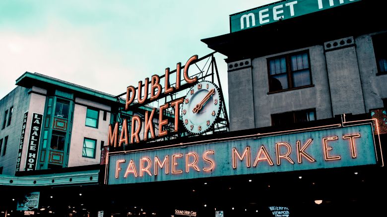 The image shows the exterior of a market with neon signs that read "Public Market" and "Farmers Market." A clock is visible between the signs.