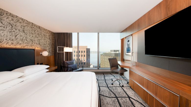 A modern hotel room with a large bed, stylish decor, TV, desk, chair, and a window offering a city view.