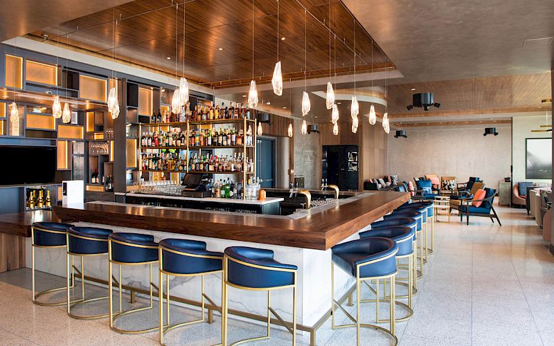 The image shows a modern bar interior with a wooden countertop, blue bar stools, pendant lights, and a fully stocked bar in the background.