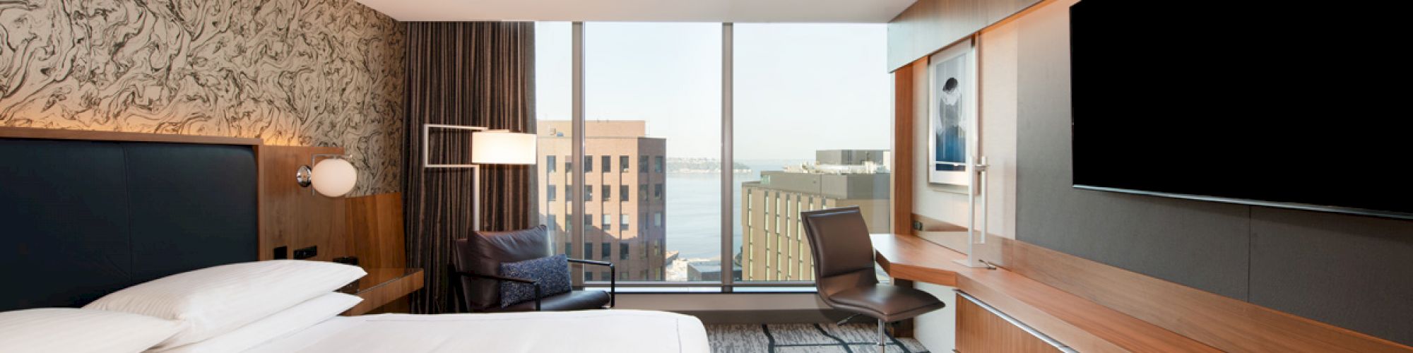 This image shows a modern hotel room with a large bed, a wall-mounted TV, a desk, and a window with a city view.