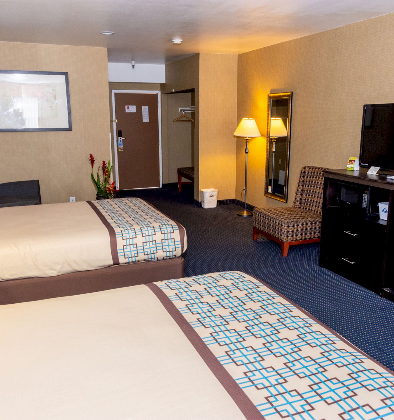 The image shows a hotel room with two beds, a TV on a stand, a lamp, a chair, wall art, a mirror, and a door in the background.