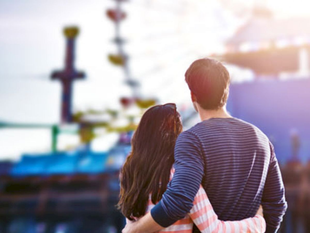 A couple stands close together, facing away from the camera, with an amusement park or fairground visible in the background.