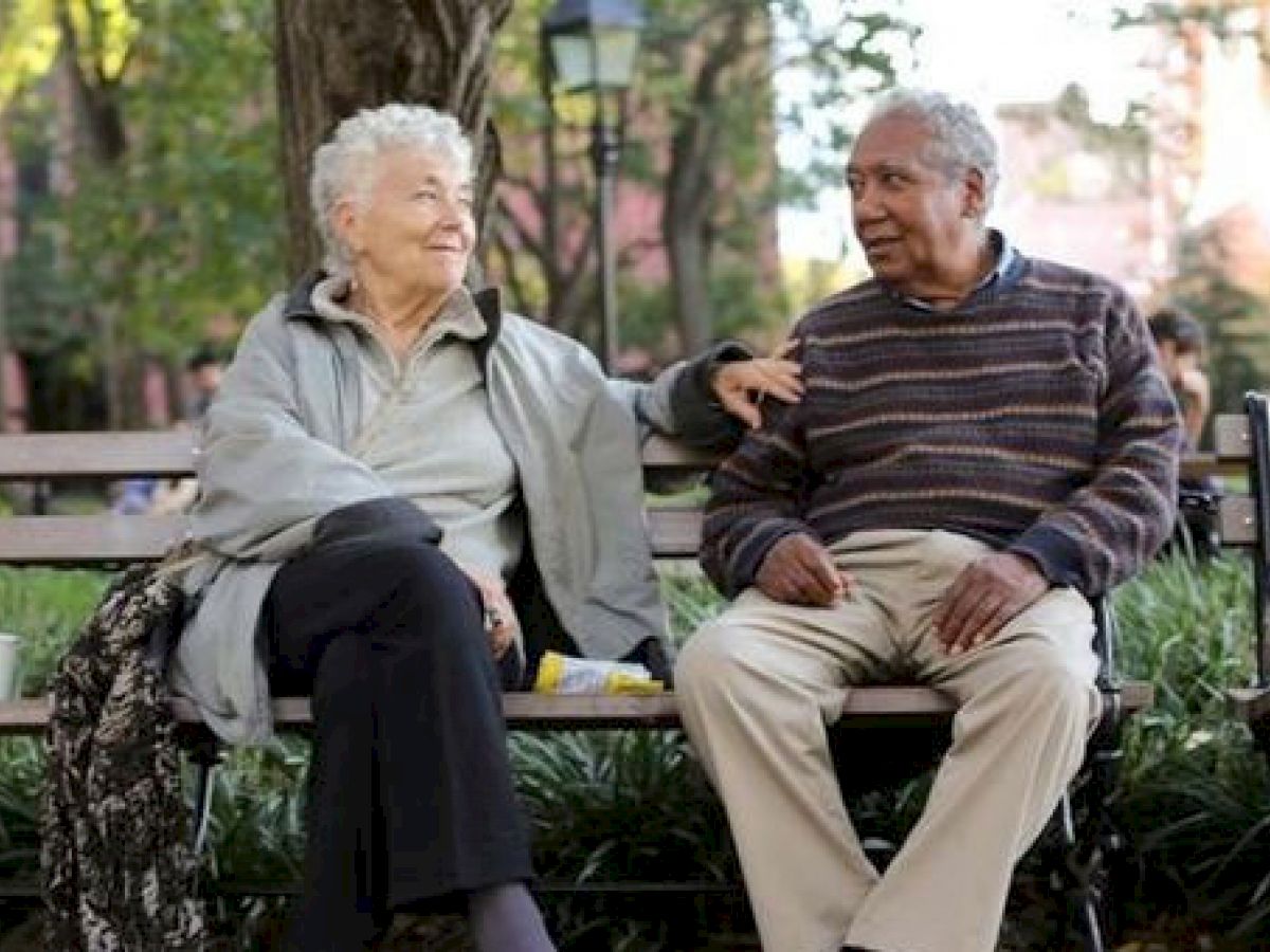 An elderly couple sits on a park bench, smiling and chatting. There are coffee cups nearby, and the scene is surrounded by greenery and trees.