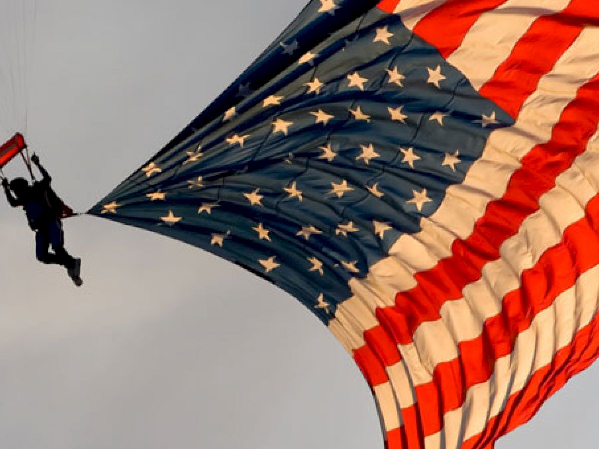 A skydiver is descending with an American flag parachute, the flag billowing dramatically in the wind against a clear sky.